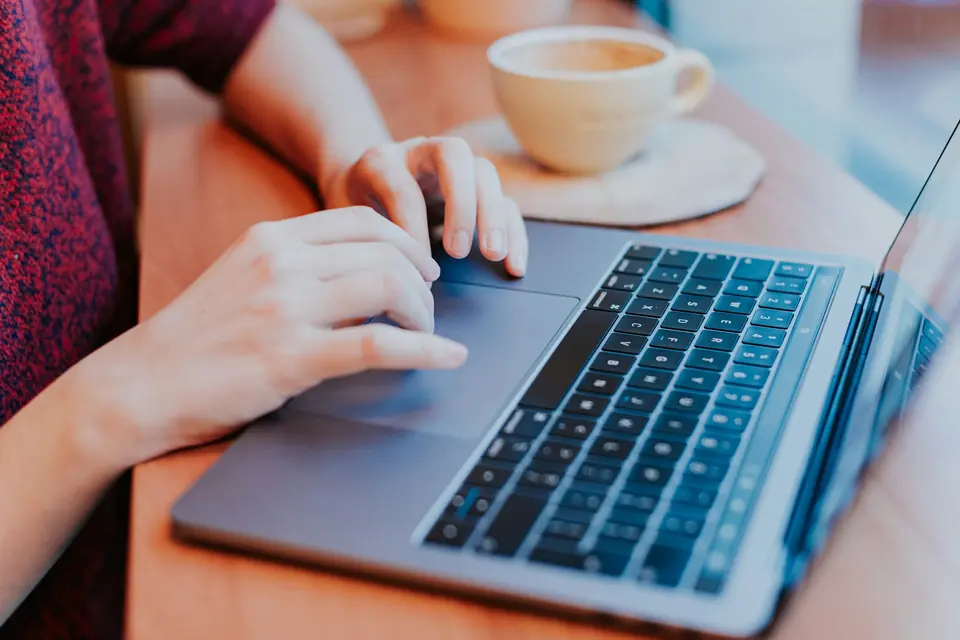 Hender på tastaturet til en bærbar PC ved siden av en kaffekopp på et trebord