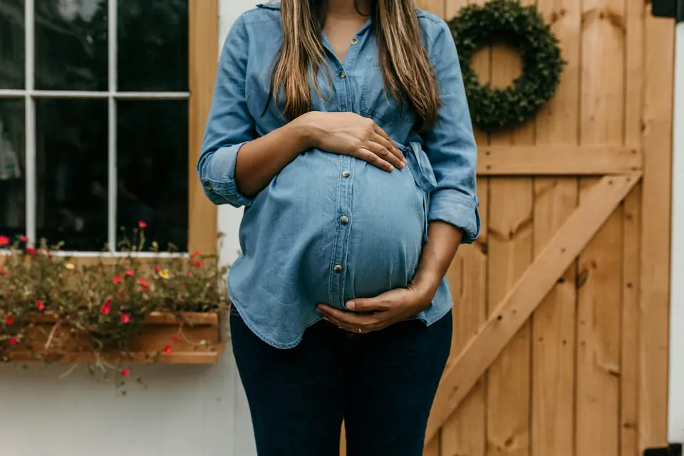 Gravid kvinne i dongeriskjorte foran en tredør og en blomsterkasse under et vindu
