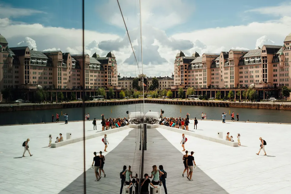 Mange mennesker på en hvit marmorplass ved vannet i Oslo, speilet i en glassvegg