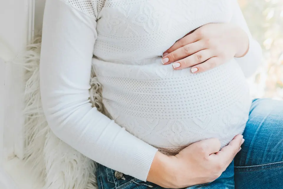Gravid kvinne i hvit genser sitter med hendene rundt magen
